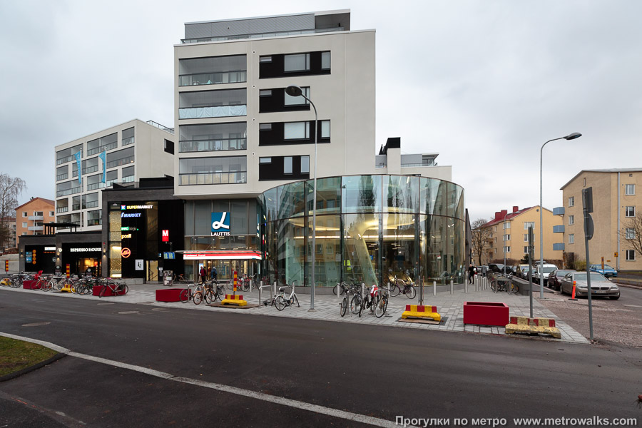 Станция Lauttasaari / Drumsö [Ла́уттасаа́ри] (Хельсинки). Наземный вестибюль станции встроен в торговый комплекс. ТЦ и дом над ним были построены вместе со строительством метро, ранее здесь была АЗС.