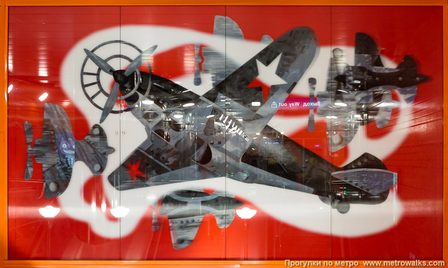 Станция Беговая (Невско-Василеостровская линия, Санкт-Петербург). Декоративное оформление стены над спуском. Над эскалаторами с противоположной платформы — картина с самолётом Савушкина на фоне падающих самолётов врагов.