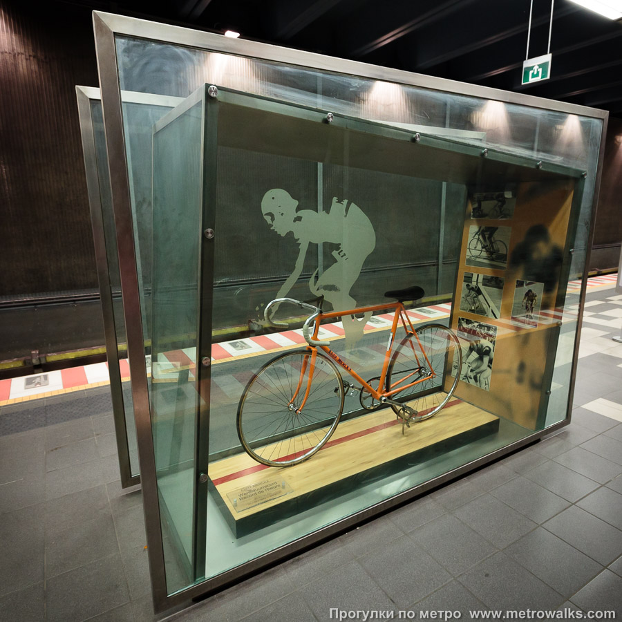 Фотография станции Eddy Merckx [Эдди Меркс] (линия 5, Брюссель). Витрина с художественной инсталляцией на платформе станции. На платформе установлена стеклянная витрина с настоящим велосипедом, на котором Эдди Меркс установил мировой рекорд «часовой гонки» в 1972 году.
