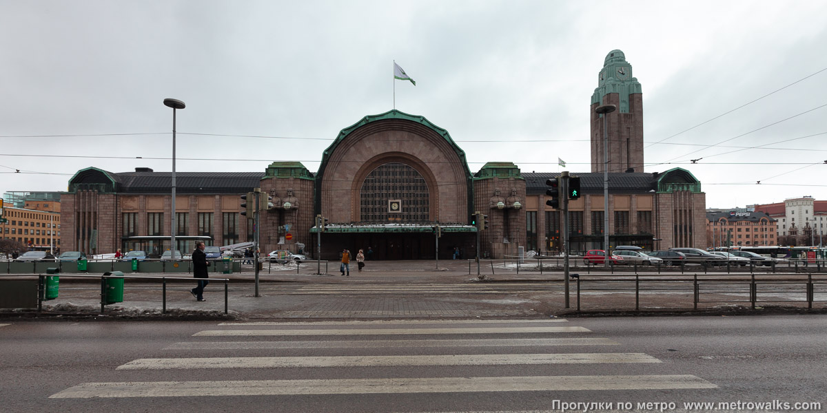 Фотография станции Rautatientori / Järnvägstorget [Ра́утатиэ́нто́ри] (Хельсинки). Вход на станцию через подземный переход, спуск в который встроен в здание. Вход в подземный вестибюль станции — через здание железнодорожного вокзала или через подземный переход на трамвайной остановке.
