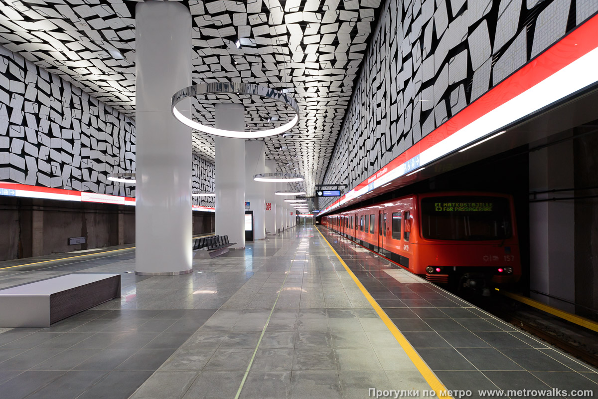 Фотография станции Urheilupuisto / Idrottsparken [У́рхейлупу́йсто] (Хельсинки). Продольный вид вдоль края платформы. Для большей цветастости — с поездом.