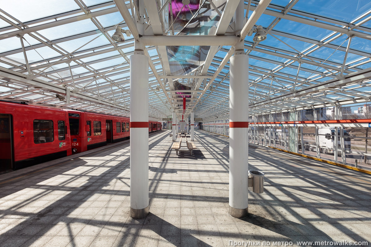 Фотография станции Vuosaari / Nordsjö [Вуо́саа́ри] (Хельсинки). Продольный вид по оси станции. Для пущей цветастости — с оранжевым поездом и голубым небом в солнечную погоду. Сверху видны панели арт-объекта «Expose».