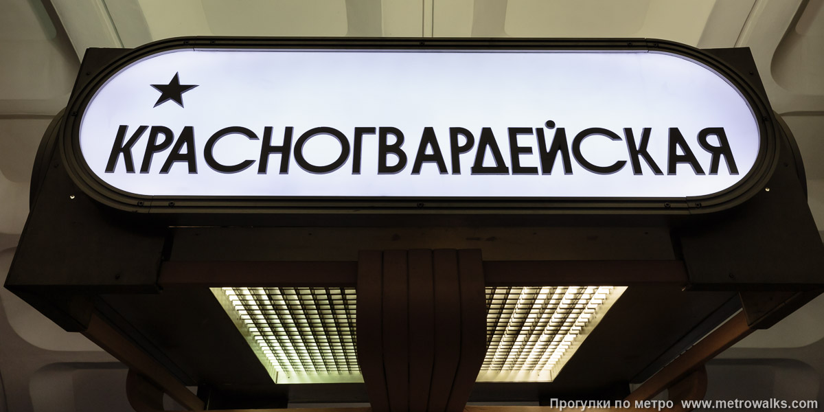 Фотография станции Красногвардейская (Замоскворецкая линия, Москва). Название станции на светильниках-торшерах, установленных внутри скамеек по оси станции. Поскольку там внутри — лампы, освещающие ВСЮ станцию, смотреть на такой указатель больно.
