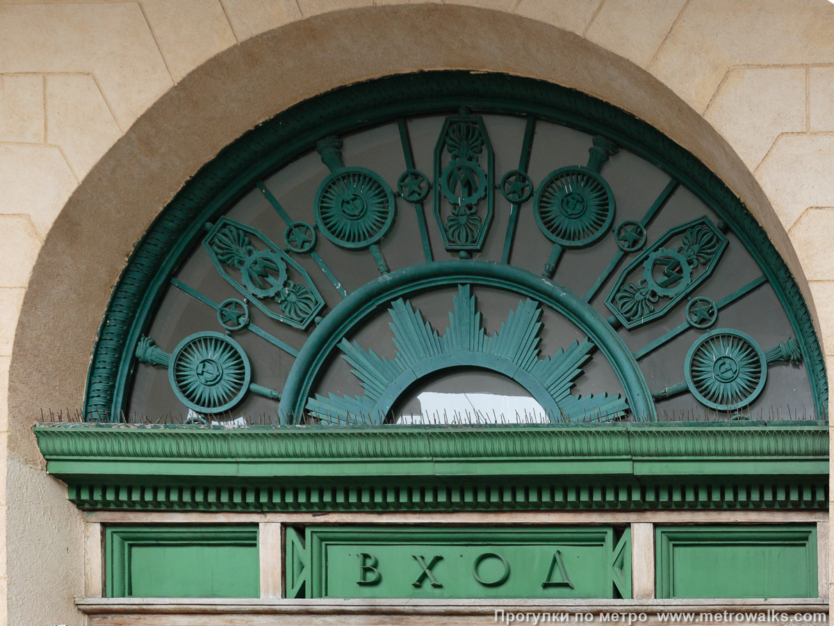 Фотография станции Кировский завод (Кировско-Выборгская линия, Санкт-Петербург). Архитектурное оформление вестибюля. Декоративная литая решётка в тимпане над входом, украшенная щитами, звёздами, серпами-и-молотами, с которыми, что необычно, сплетается логотип метрополитена в виде буквы «М».