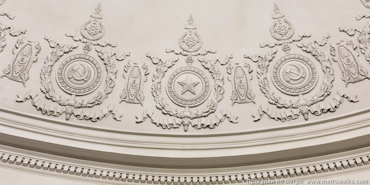 Фотография станции Нарвская (Кировско-Выборгская линия, Санкт-Петербург). Потолок эскалаторного зала. Лепнина со звёздами на щитах и серпами-и-молотами в растительном орнаменте — типовые советские символы Красной армии и рабоче-крестьянского труда.
