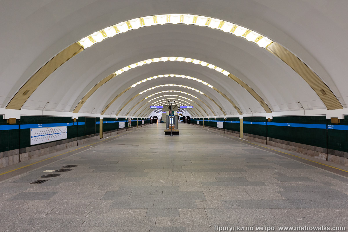 Фотография станции Удельная (Московско-Петроградская линия, Санкт-Петербург). Общий вид по оси станции от входа в сторону глухого торца. Нагромождение лишних объектов на платформе полностью закрывает изначальную перспективу мемориала в дальнем торце.
