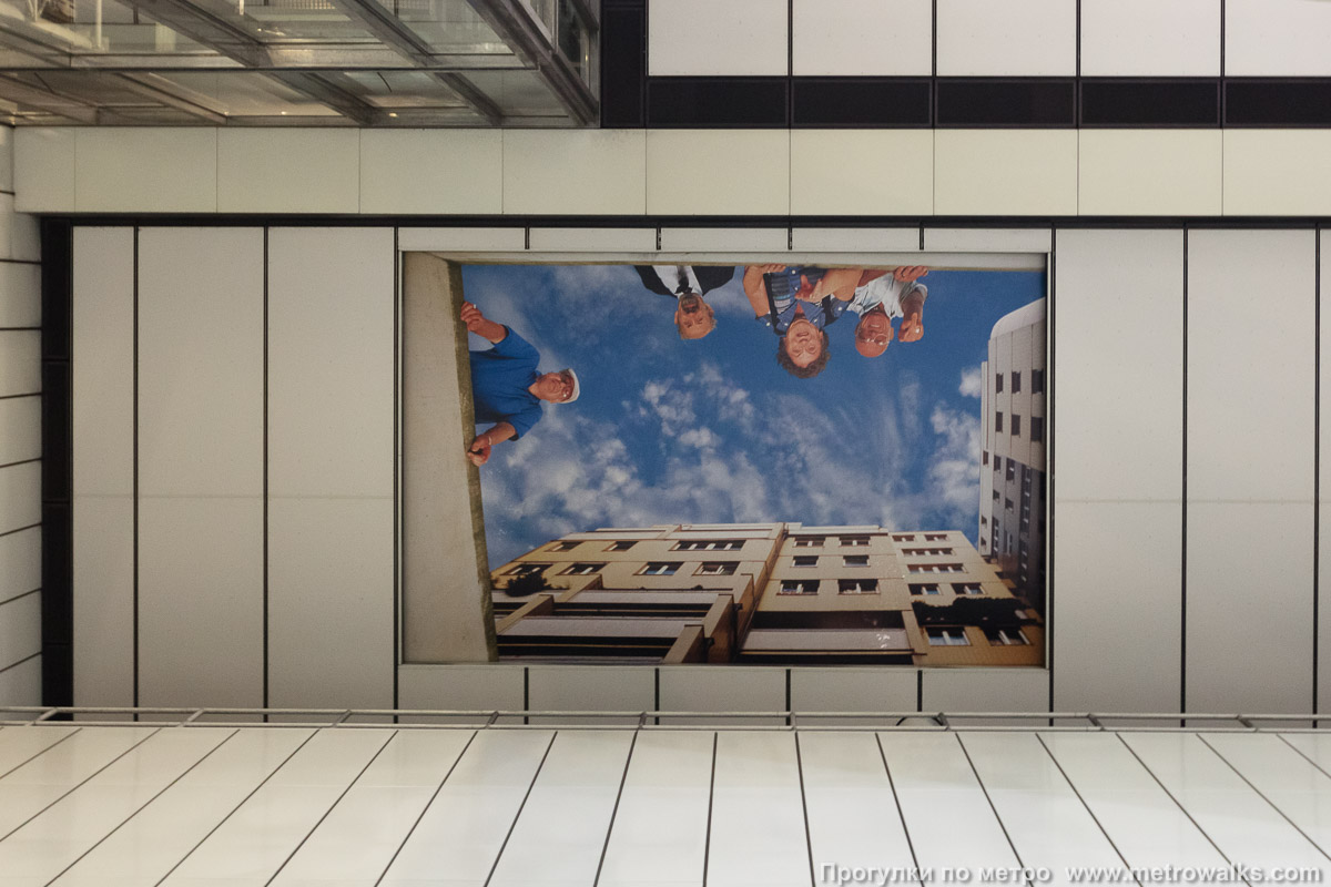 Фотография станции Enkplatz [Энкплац] (U3, Вена). Декоративная отделка потолка в проходе к эскалаторам. Потолок аванзала украшает картина, изображающая окно на улицу: видно небо, дома рядом со станцией и местные жители, с интересом смотрящие с поверхности вниз на станцию. Художники — Ilse Haider и Mona Hahn.