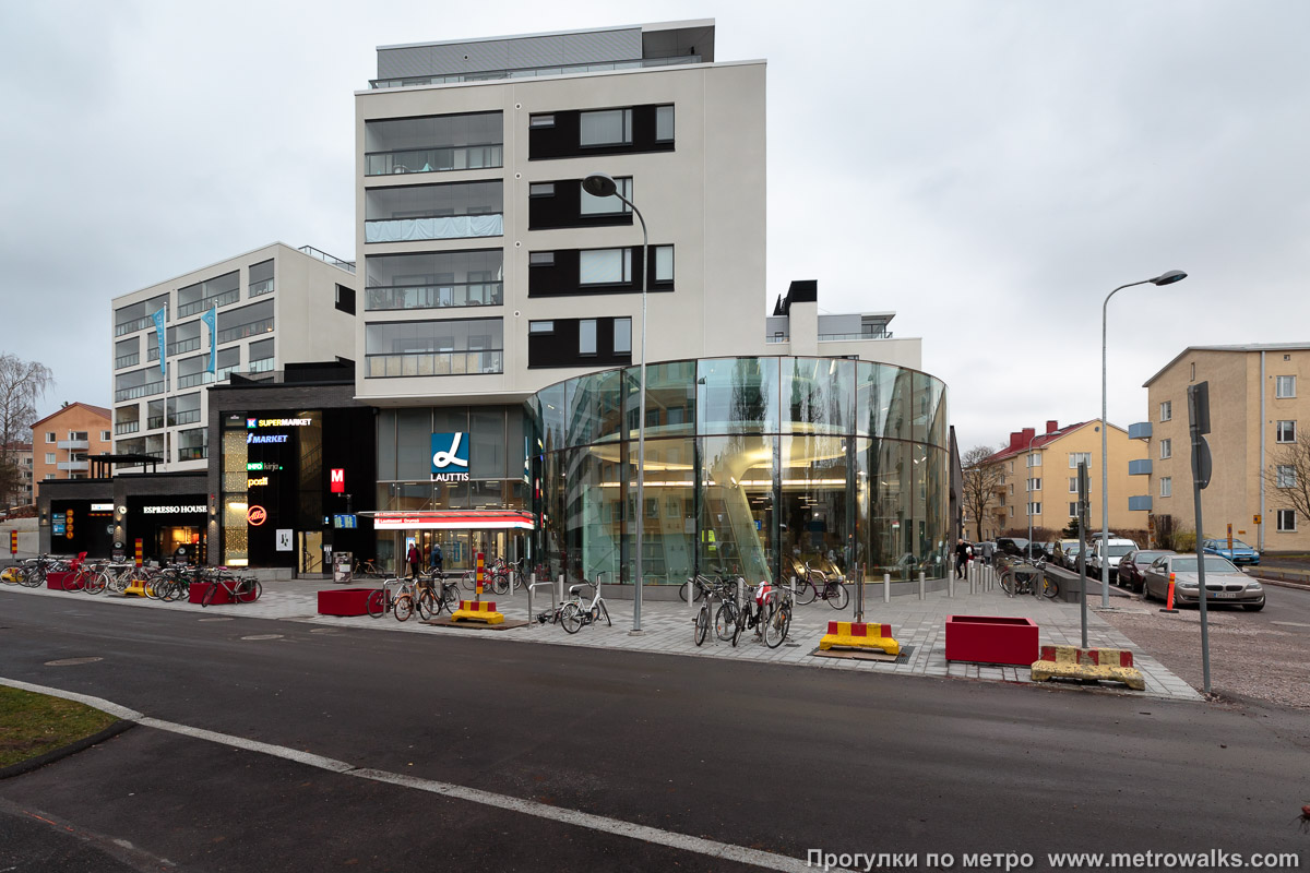 Фотография станции Lauttasaari / Drumsö [Ла́уттасаа́ри] (Хельсинки). Наземный вестибюль станции встроен в торговый комплекс. ТЦ и дом над ним были построены вместе со строительством метро, ранее здесь была АЗС.