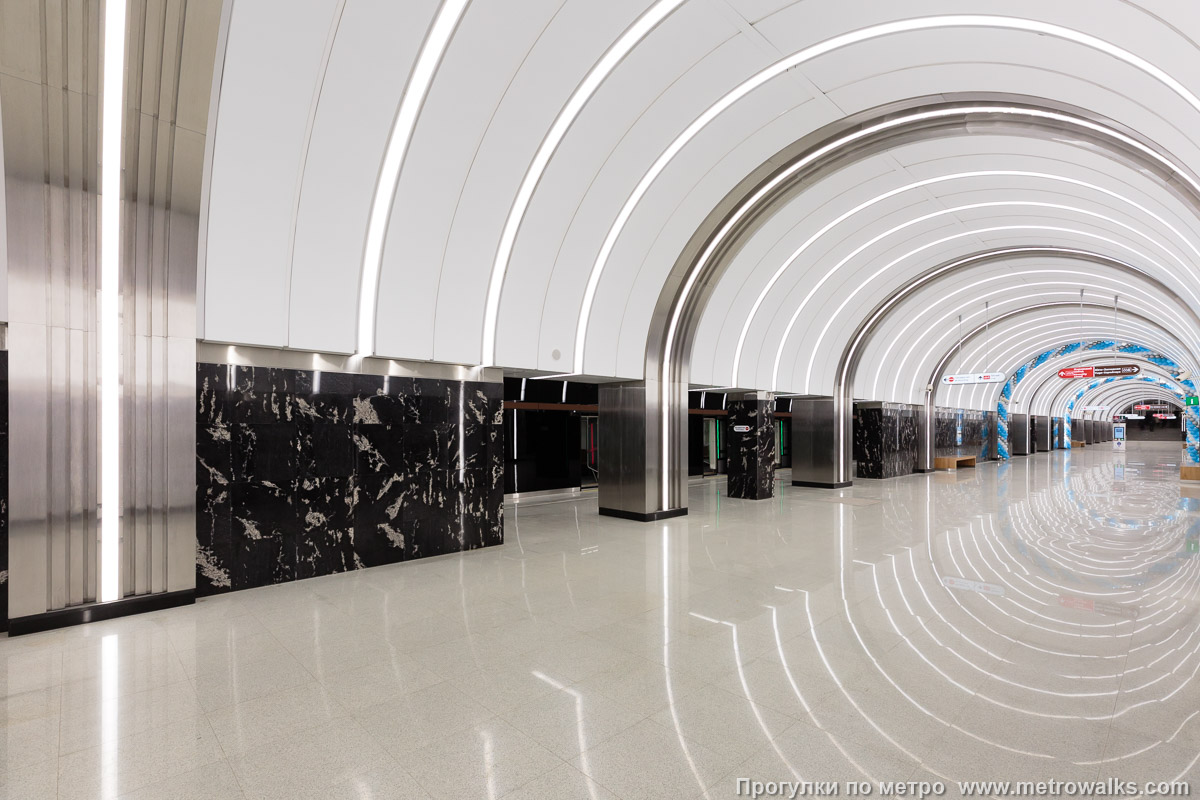 Фотография станции Путиловская (Красносельско-Калининская линия, Санкт-Петербург). Вид по диагонали. Световые арки, спускающиеся по колоннам до самого пола, выглядят ослепительно и создают неудобство пассажирам даже с небольшими недостатками зрения.