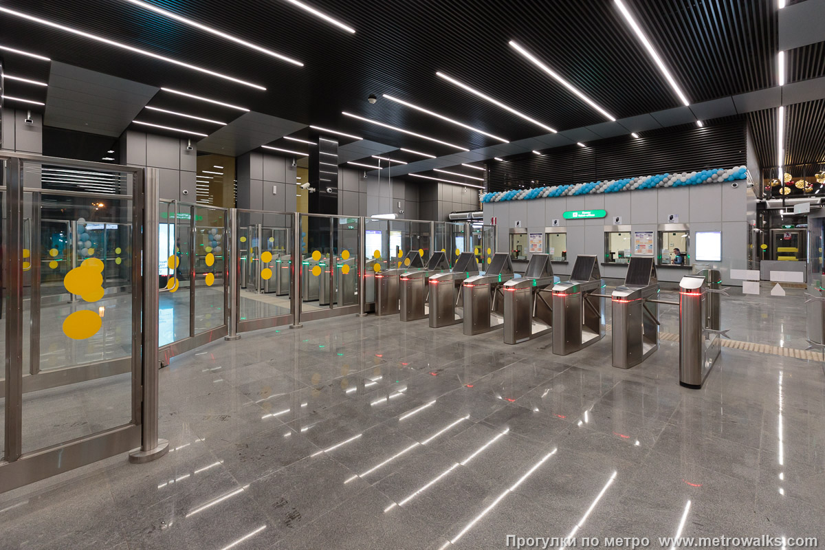 Фотография станции Путиловская (Красносельско-Калининская линия, Санкт-Петербург). Внутри вестибюля станции, общий вид. От входных эскалаторов. При наличии турникетов в обоих направлениях непонятен смысл барьера как до турникетов, так и после них. На станции Кировский завод в аналогичной конфигурации его нет.