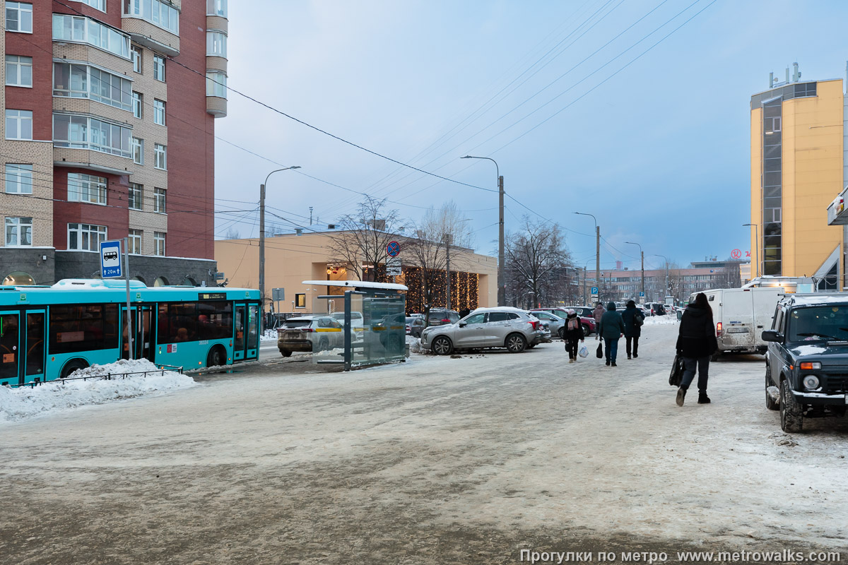 Фотография станции Путиловская (Красносельско-Калининская линия, Санкт-Петербург). Общий вид окрестностей станции. Улица Васи Алексеева от улицы Маршала Говорова со стихийной парковкой на тротуаре. На заднем плане — Кировский завод.