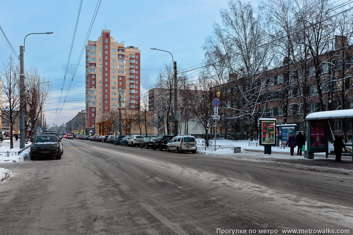 Фотография станции Путиловская (Красносельско-Калининская линия, Санкт-Петербург). Общий вид окрестностей станции. Улица Васи Алексеева от проспекта Стачек и Кировского завода. Непонятно, почему не поменяли местами автобусную остановку и парковку вдоль края проезжей части у метро.