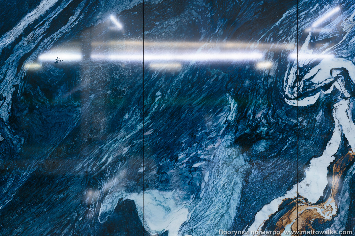 Фотография станции Зенит (Невско-Василеостровская линия, Санкт-Петербург). Декоративная отделка стены в проходе к эскалаторам. Фрагменты стен облицованы искусственным камнем тёмно-синего цвета с белыми разводами, изображающими пенящиеся волны Финского залива.